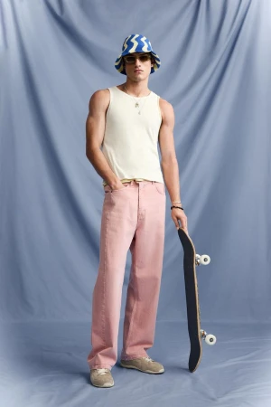 Full shot of a young adult male in a tank top and light pink jeans holding a skateboard in front of a blue backdrop.