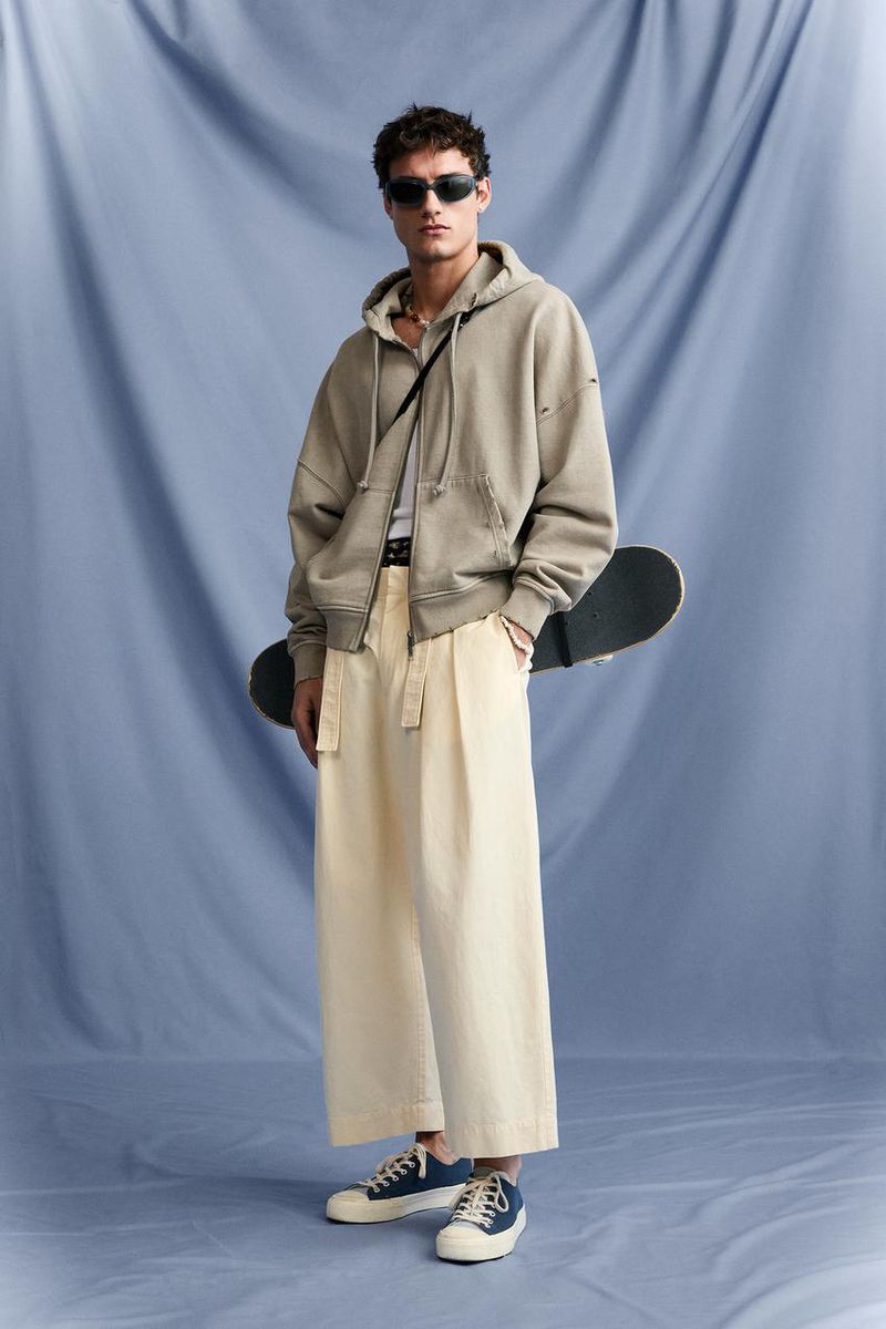 Young man in a hooded sweatshirt and baggy pants poses with a skateboard against a blue muslin backdrop in a fashion studio setting.