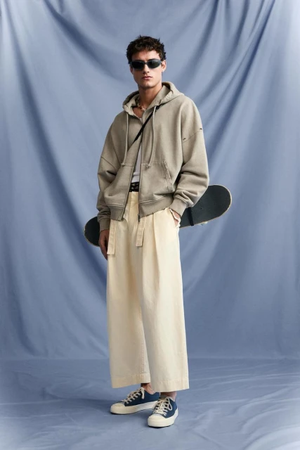Young man in a hooded sweatshirt and baggy pants poses with a skateboard against a blue muslin backdrop in a fashion studio setting.