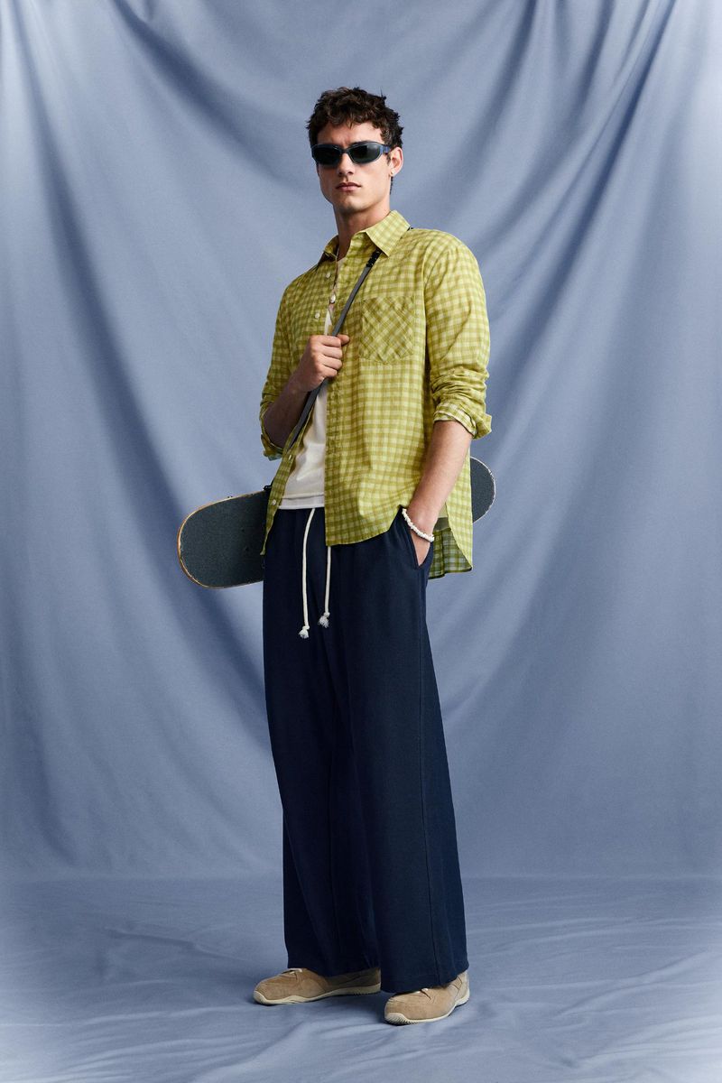 Full-length portrait of a male model wearing sunglasses, yellow patterned shirt, dark trousers, and holding a skateboard