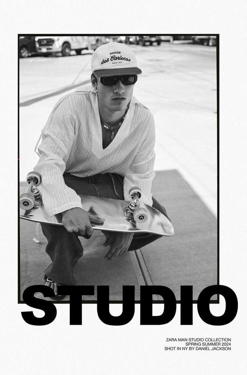 Black and white image of a crouched male model with a skateboard on a street. He is wearing sunglasses and a baseball cap.