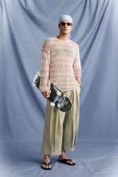 Model wearing a pastel knit sweater, pleated trousers, sunglasses, and a headscarf holding a skateboard against a grey backdrop