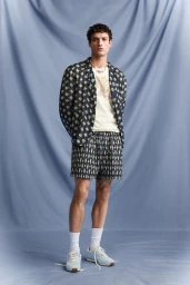 Male model wearing patterned blazer and matching shorts with white socks and sneakers against a draped blue backdrop.