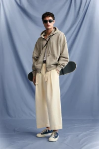 Male model wearing a beige hoodie and light trousers holding a skateboard against a draped blue backdrop