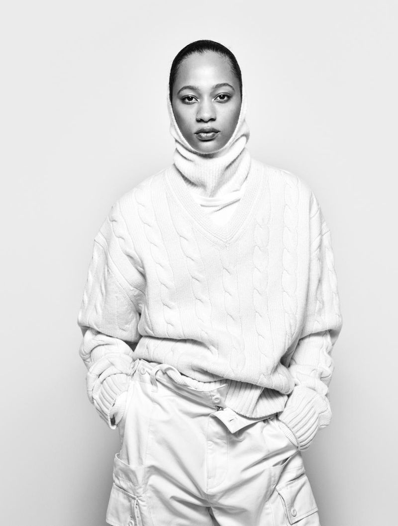 Model wearing a white cable-knit sweater and turtleneck against a seamless light grey background