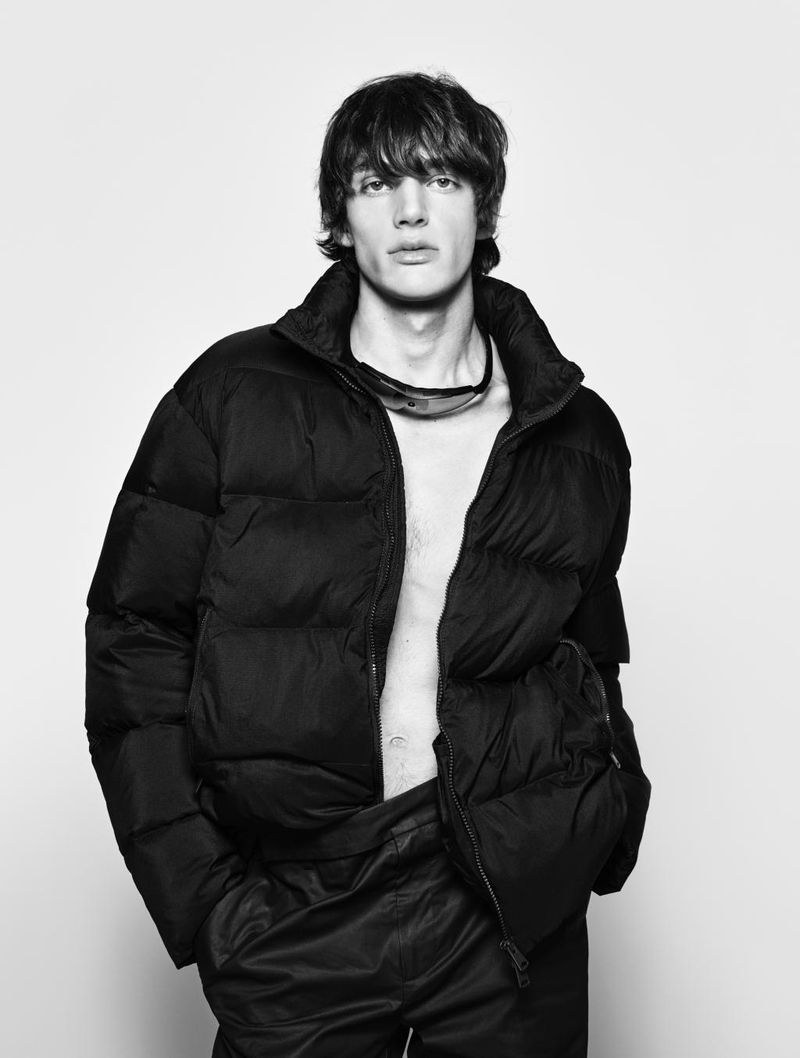 Monochrome studio portrait of a male model wearing a puffy jacket with sunglasses around his neck, against a neutral backdrop.
