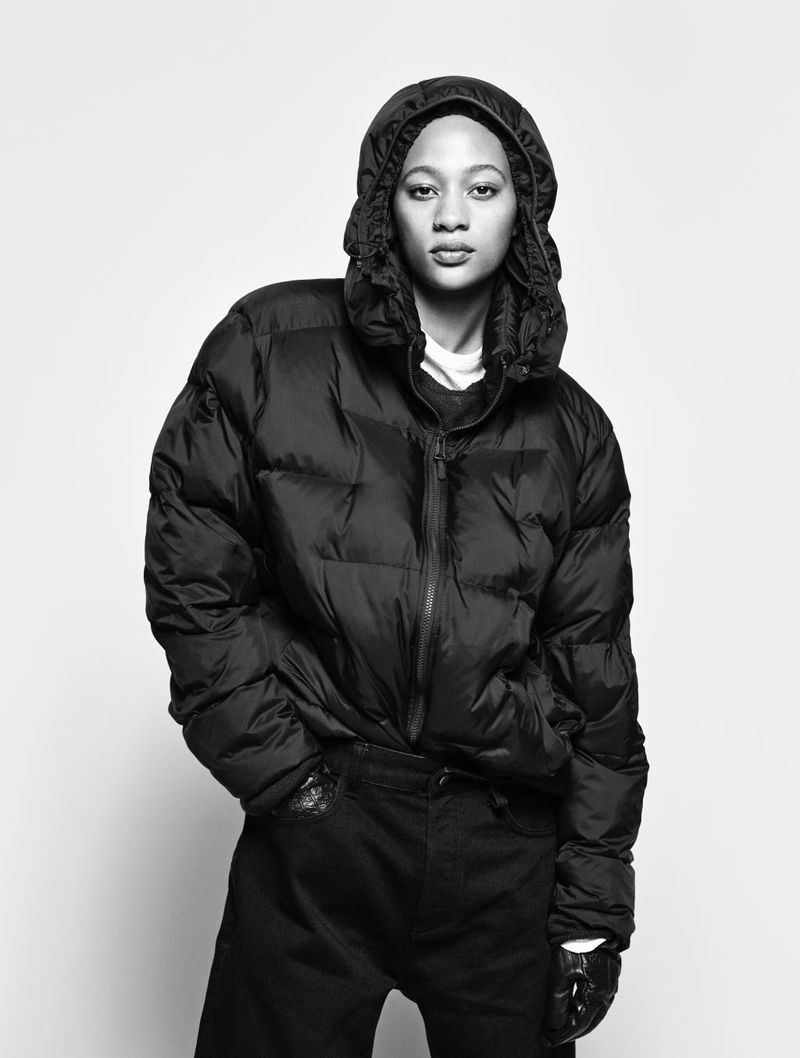 Black-and-white portrait of a young adult woman in a hooded black puffer jacket and black jeans, posing against a plain white backdrop.