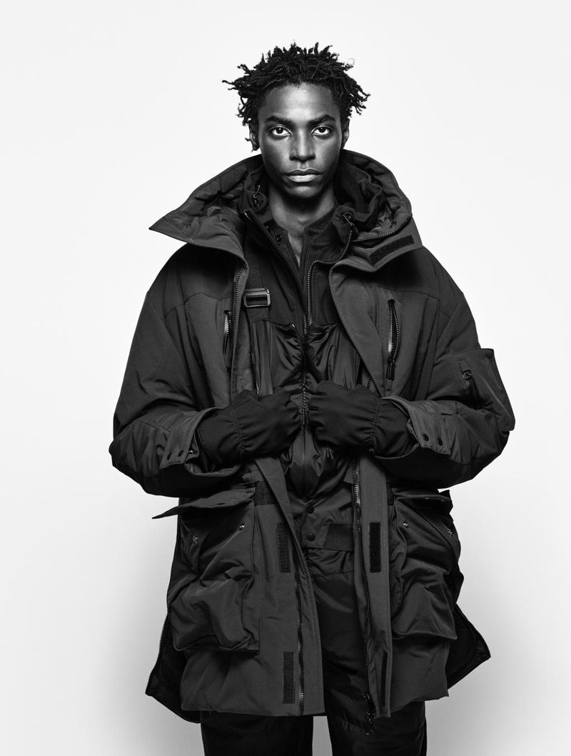 Monochromatic portrait of a Black male model wearing a parka coat against a bright white backdrop in a studio setting. Fashion editorial.