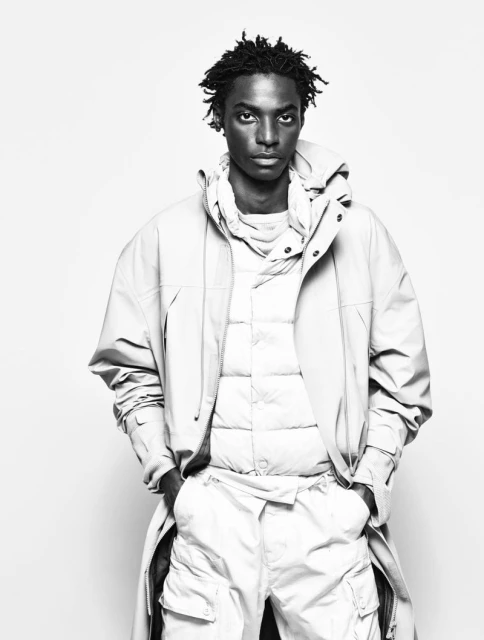 Black male fashion model poses in a hooded coat, quilted vest, and cargo pants against a white background in a studio shot.