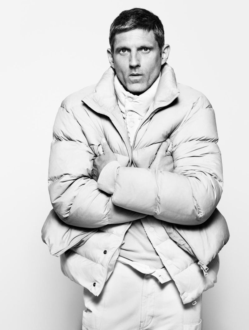 Monochrome portrait of a man in a puffer jacket with arms crossed, against a stark white backdrop, showcasing fashion and style.