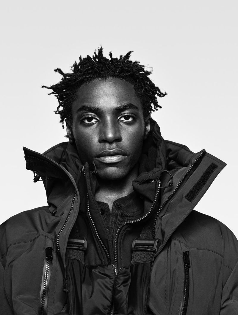 Black and white portrait of an African-American man with dreadlocks wearing a black down jacket with a hood against a neutral background.