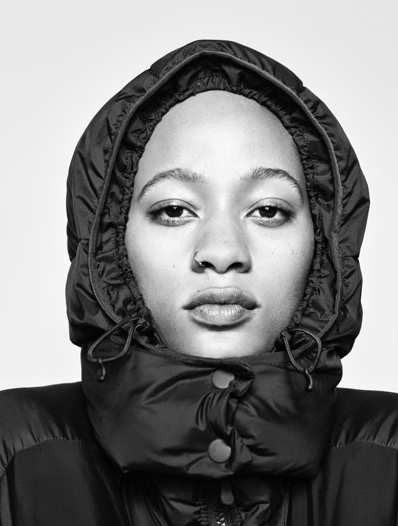 Close-up portrait of a young adult woman with light skin, wearing a black hooded puffer jacket, against a neutral gray backdrop.