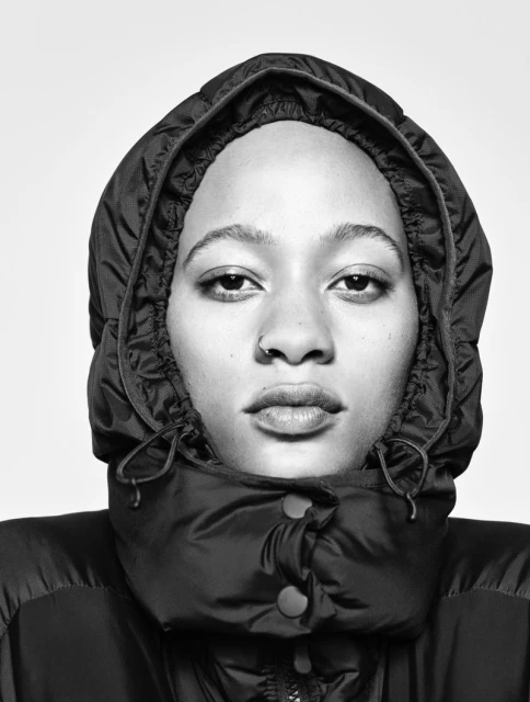 Close-up portrait of a young adult woman with light skin, wearing a black hooded puffer jacket, against a neutral gray backdrop.
