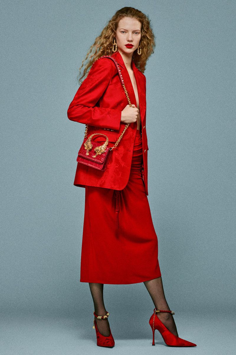 Fashion shot: Woman in a red blazer and skirt with a red purse over her shoulder, red heels, and patterned hose against a plain background.