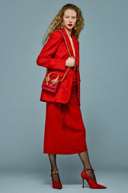 Fashion shot: Woman in a red blazer and skirt with a red purse over her shoulder, red heels, and patterned hose against a plain background.