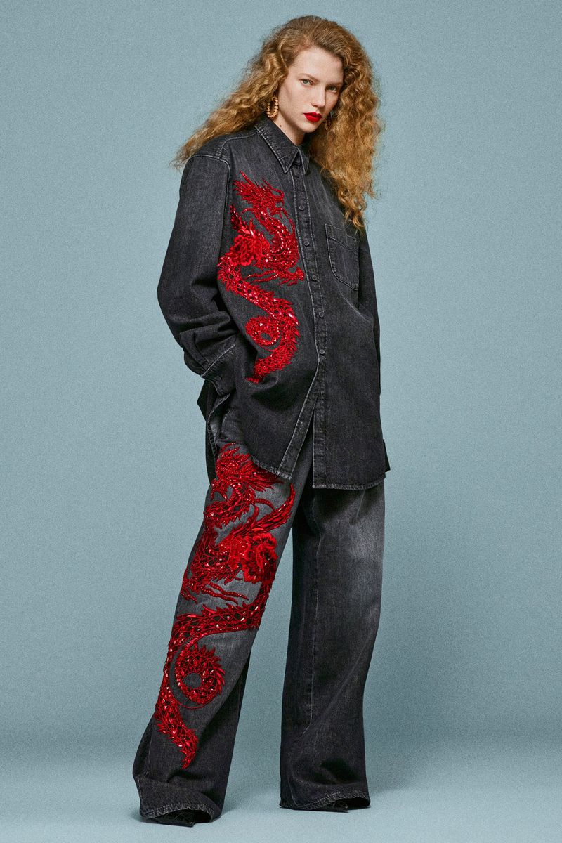 A redhead fashion model in a denim shirt and pants with red dragon designs poses against a light blue backdrop in a studio setting.