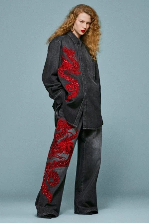 A redhead fashion model in a denim shirt and pants with red dragon designs poses against a light blue backdrop in a studio setting.