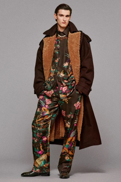 A man poses in a floral-print shirt and pants, with a brown coat featuring a sherpa lining, against a gray studio backdrop.