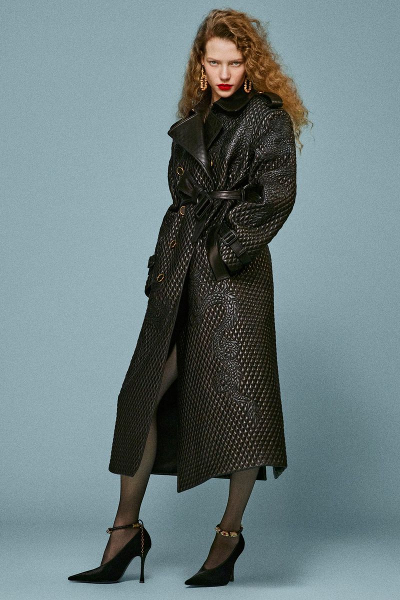 A curly-haired woman in a patterned black trench coat, matching heels, and fishnet stockings poses against a muted blue background.