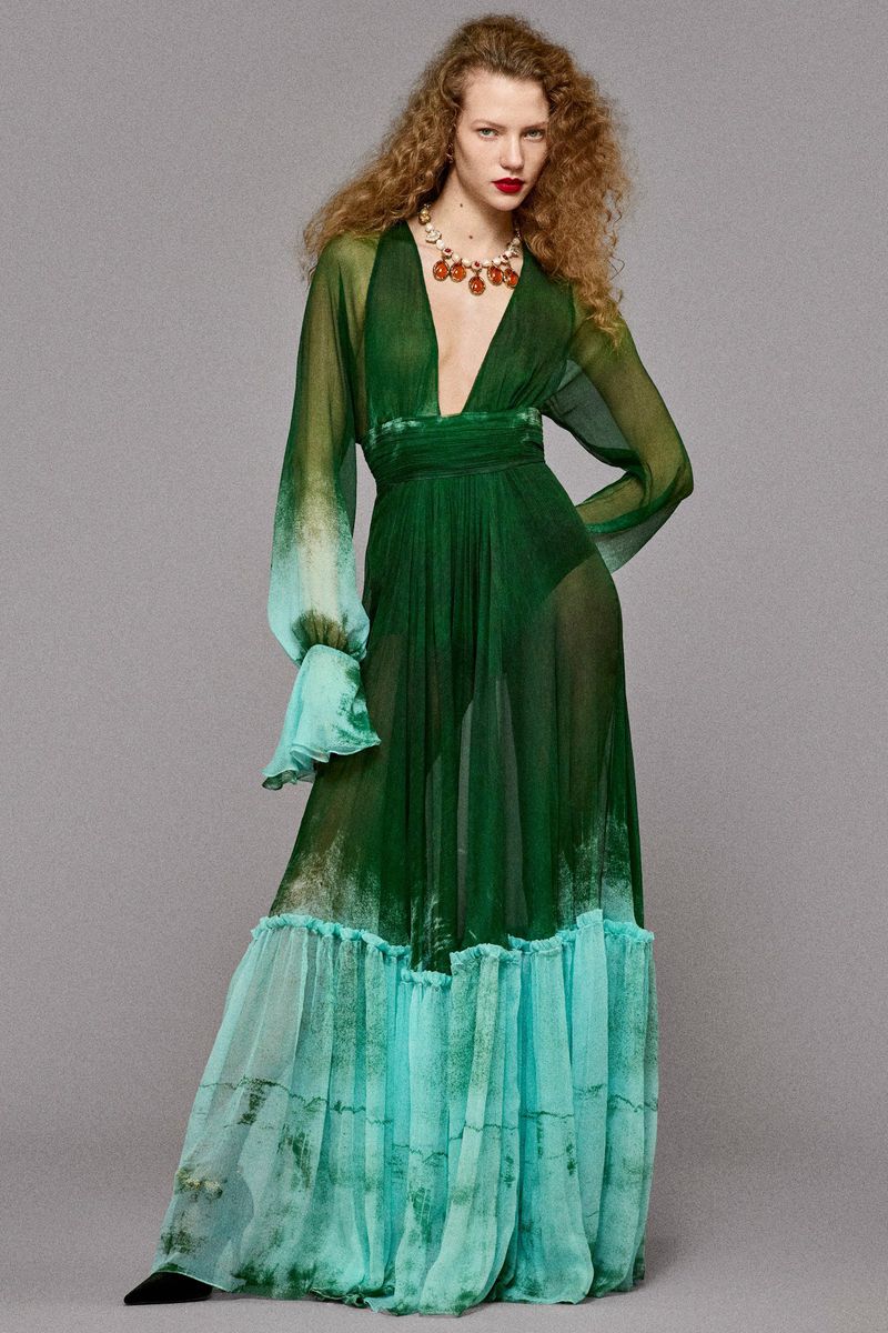 Full-length portrait of an adult woman in a flowing green gown with an ombre effect, wearing a statement necklace against a grey background.