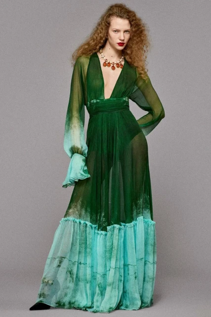 Full-length portrait of an adult woman in a flowing green gown with an ombre effect, wearing a statement necklace against a grey background.
