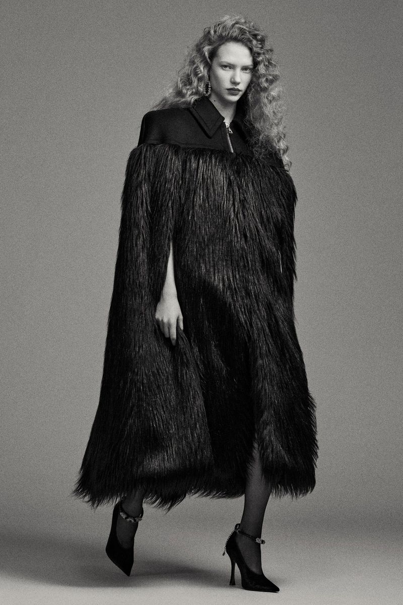 Model with long curly hair wearing a textured black cape-like coat and heels against a grey studio background