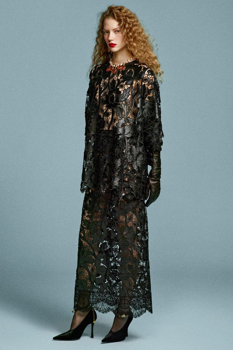 Woman in a black lace dress and gloves stands in a photo studio. She is wearing high heels and a red necklace. The background is light blue.