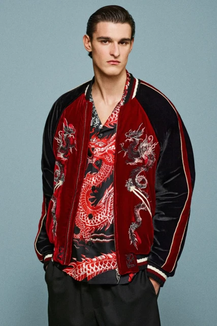 A man in a red velvet jacket with dragon embroidery and a matching shirt, posing against a pale blue studio backdrop.