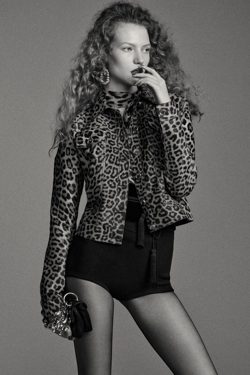 Model with long curly hair wearing a leopard print jacket and dark shorts against a grey backdrop