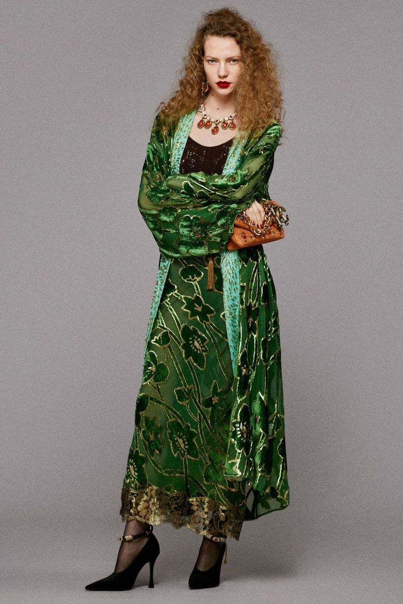 Full-length shot of a young woman with curly hair wearing a green floral dress, long floral coat, and high-heeled shoes against a grey background.