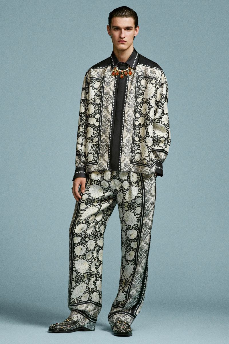 Full-length shot of a male model wearing a patterned shirt and pants, accessorized with a necklace, against a light blue background.