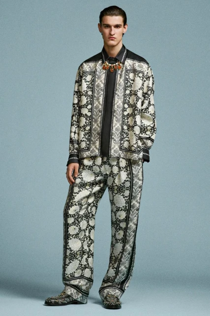 Full-length shot of a male model wearing a patterned shirt and pants, accessorized with a necklace, against a light blue background.