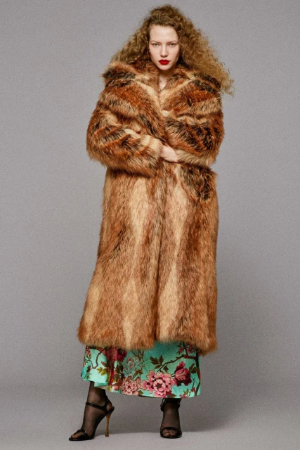 Woman with curly hair wears a long fur coat over a floral dress, black heels, posing against a neutral backdrop in studio lighting.