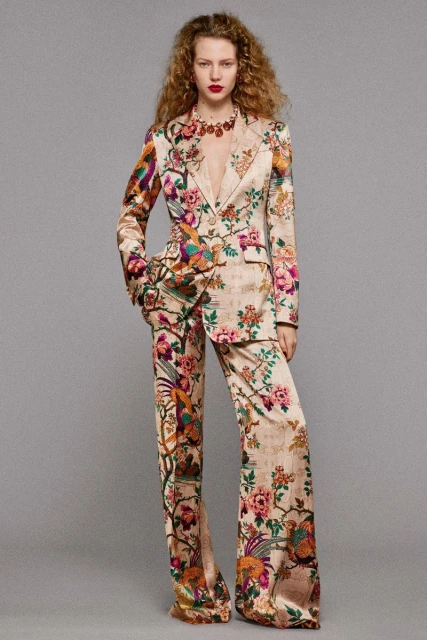 Adult woman wearing a floral patterned satin pantsuit and statement necklace in a studio setting with a neutral gray background.