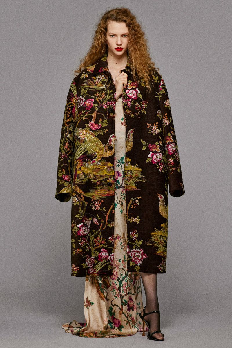 A woman wearing a long, patterned brown coat over a floral dress and black high heels, posing in front of a neutral gray background.