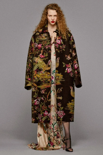 A woman wearing a long, patterned brown coat over a floral dress and black high heels, posing in front of a neutral gray background.
