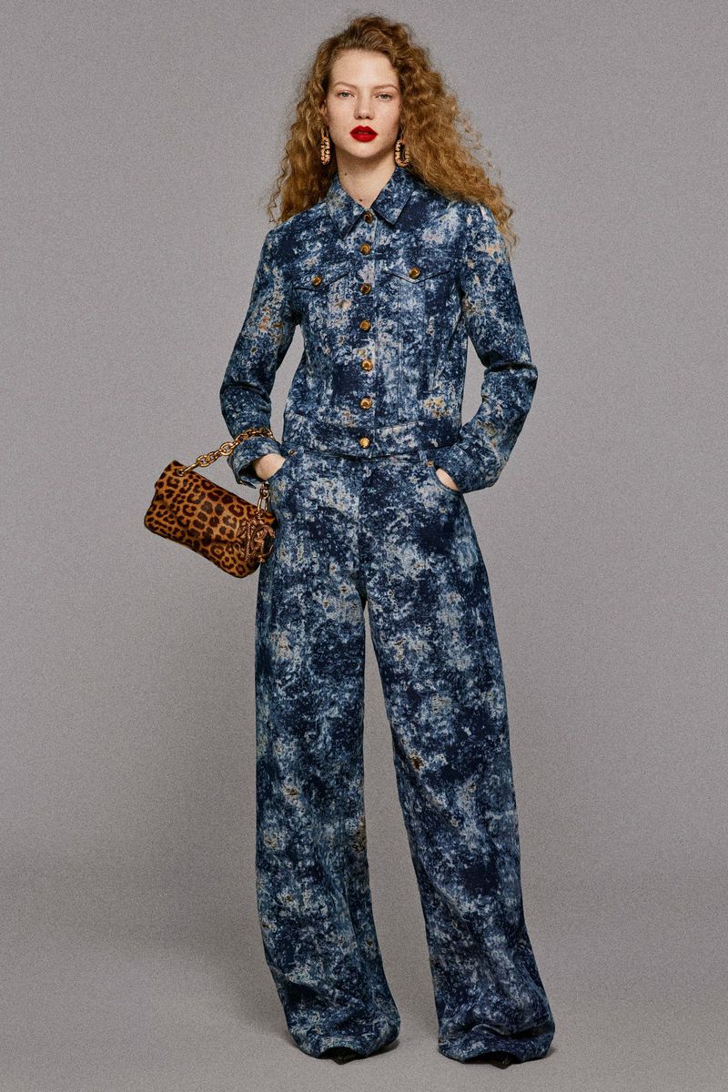 A fashionable adult female with curly hair is posing in an all-over printed blue denim jacket and pants with a patterned handbag.