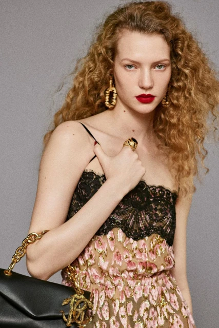 Fashion portrait of a curly-haired model wearing black lace and a gold dress with gold earrings, rings and a leather purse.