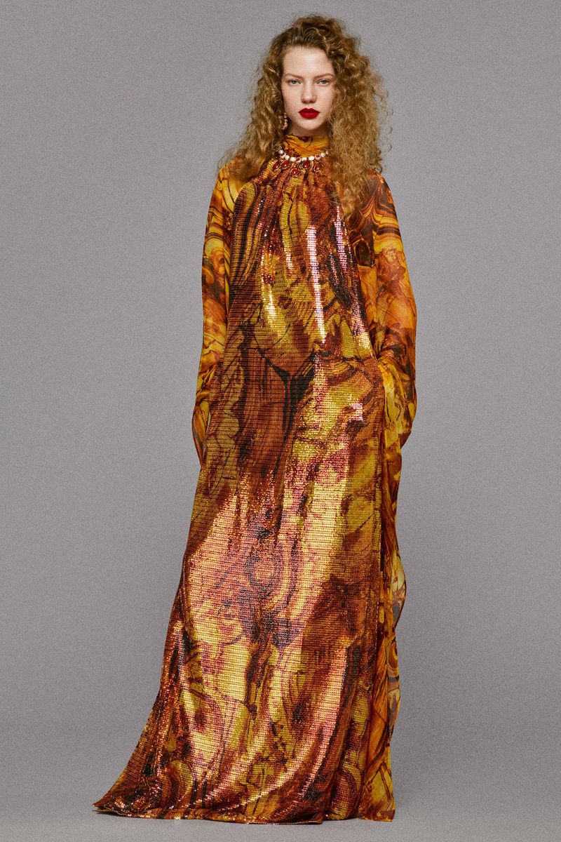 A woman in a long, patterned dress with curly hair, looking at the camera against a neutral background, showcasing fashion and style.