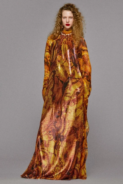 A woman in a long, patterned dress with curly hair, looking at the camera against a neutral background, showcasing fashion and style.