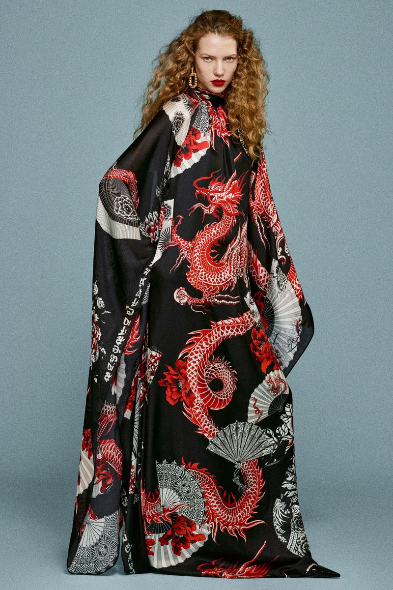 A woman with curly auburn hair models a black silk dress with a vibrant red dragon design against a serene blue backdrop.