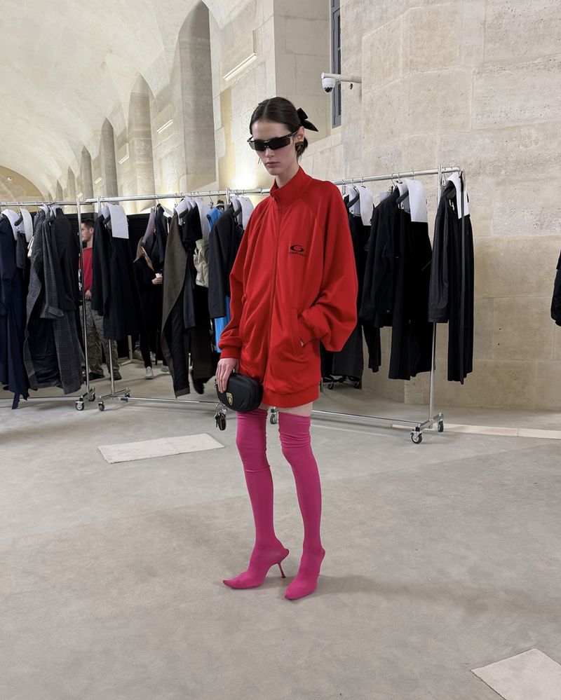A fashion model poses indoors wearing a red tracksuit, pink thigh-high boots, sunglasses, and a black purse, in front of a rack of clothes.