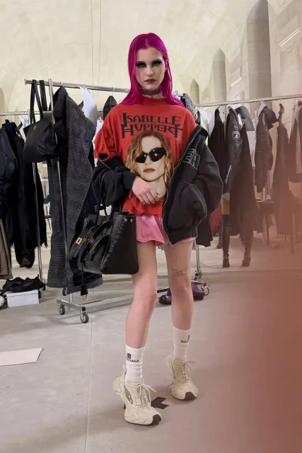 Young woman with pink hair wearing a red graphic tee, pink skirt, bomber jacket, and sneakers, standing in an indoor setting with clothes rack.