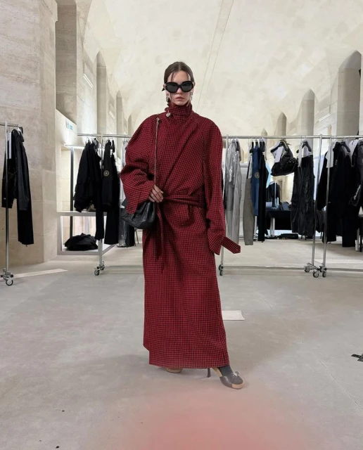A woman in a red-and-black checkered coat stands in an indoor location. She wears sunglasses, has a black purse, and is looking at the camera.