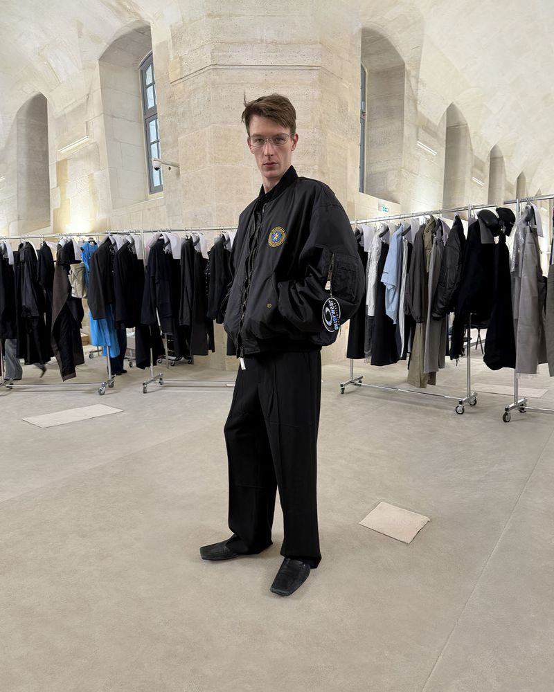 Adult male in glasses poses in front of rack of clothing in a large room with arched ceilings, wearing a black bomber jacket and pants.