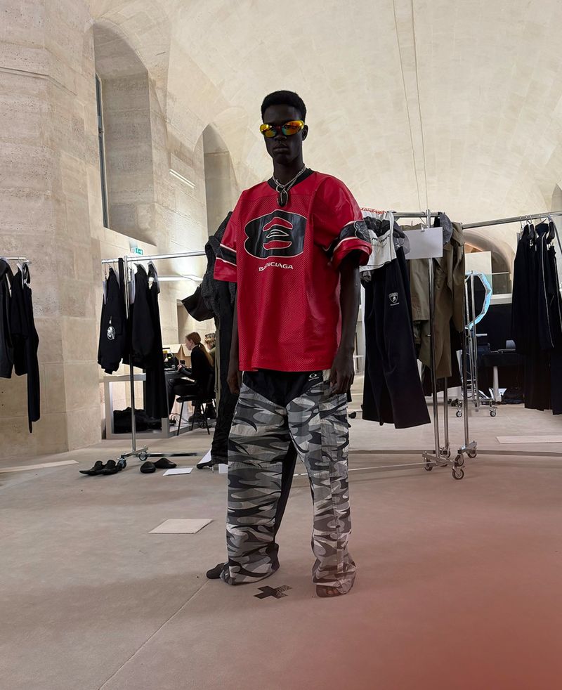 A person stands in a red Balenciaga shirt and camouflage pants, posing inside a garment storage and prep area.