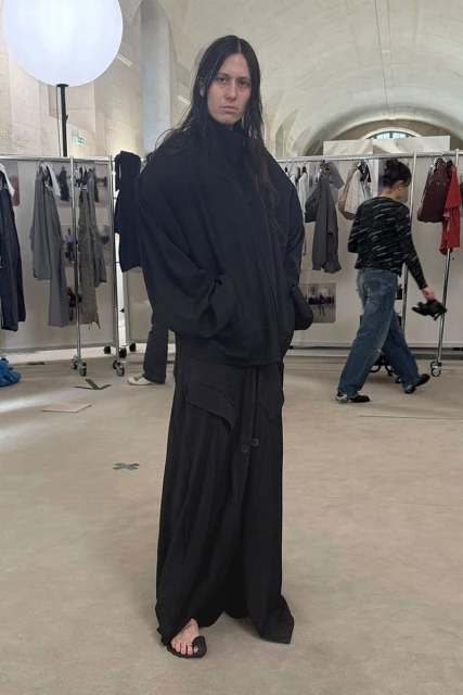 Adult female fashion model wearing an oversized black hoodie and long black skirt standing indoors during a fashion show backstage.