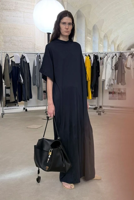 A woman in a black dress and sandals carries a large black leather bag with a lock. Background features clothing racks and architectural details.
