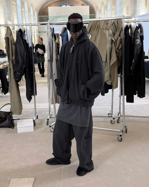 Male fashion model poses in oversized black clothing with futuristic visor glasses, surrounded by racks of clothing in an indoor setting.