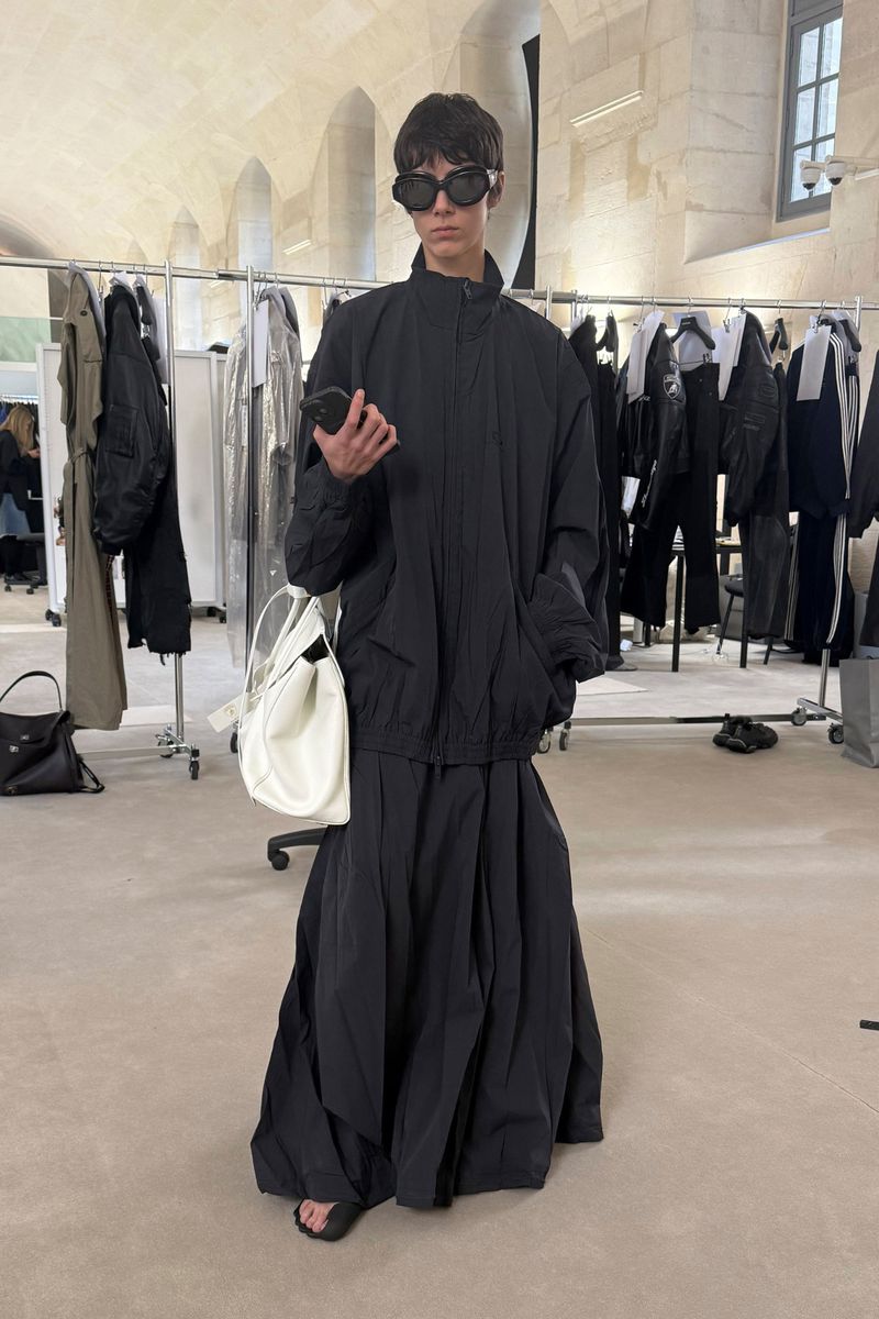 Model in black jacket and skirt wearing sunglasses and holding a white handbag in a showroom
